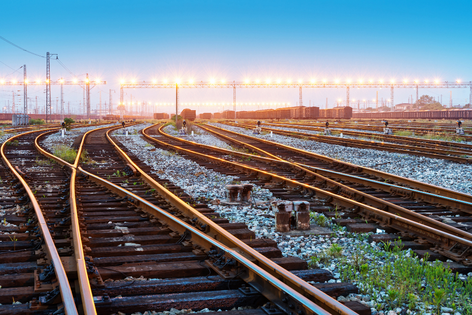Several sets of railroad tracks merge and cross and disappear into the background, which shows a train yard in the far background that is lit by a row of bright lights.