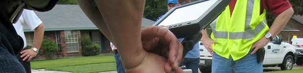 Close-up of a pair of hands holding and interacting with a screen. In the background, several men, mostly at torso level, appear to be observing. One of them is wearing a safety-yellow reflective vest and there is a truck in the background behind him.