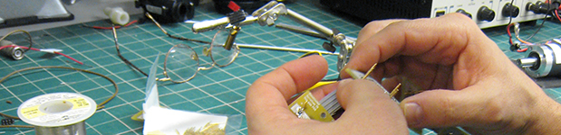 A close-up of hands assembling a small electrical product over a busy workspace. The green, gridded mat on the workspace has various spools of wire, a pair of glasses, and assorted other tools and machinery.