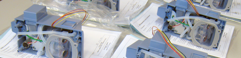 A close-up of several grayish blue plastic prototypes with clear tubing at the back and colorful wires sticking out, each sitting over a sheaf of paper documentation.