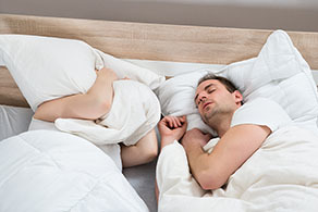 A couple is shown in bed, with one struggling to sleep, with a pillow over their face, while the other snores.