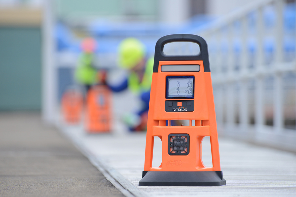 The Radius on a job site. It resembles an orange traffic cone but has an obvious display and a black handle and black base. Two more devices are seen being set up in the background.