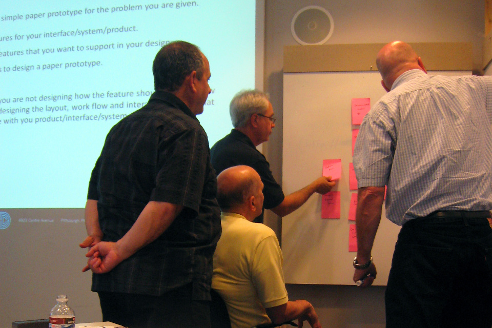 Four gentlemen crowd around a whiteboard as they affinity sort sticky notes.