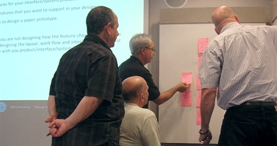 Four gentlemen crowd around a whiteboard as they affinity sort sticky notes.
