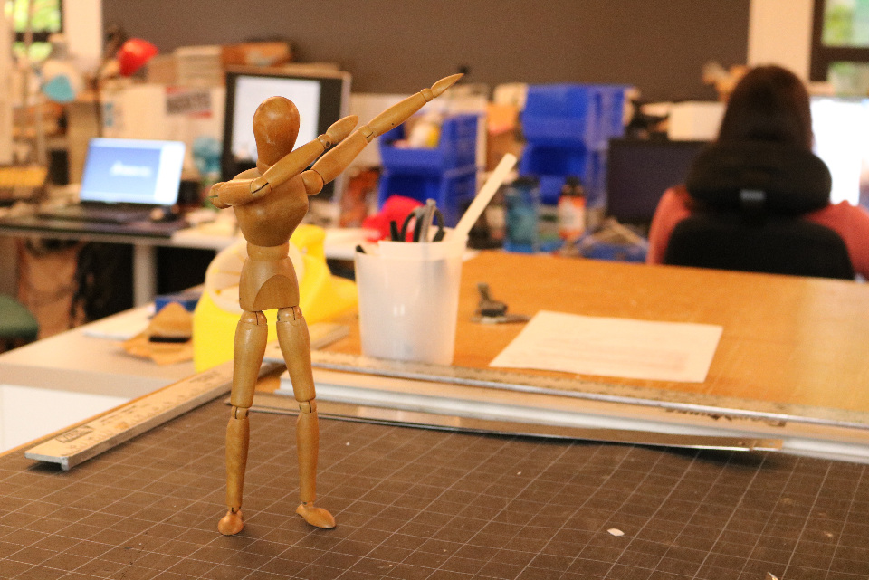 An articulated small wooden model of a human body stands on a table while people on computers can be seen in the background.