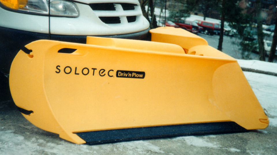 A yellow Solotec snow plow is shown attached to a white truck on a city street.