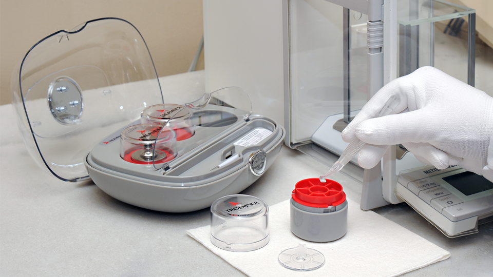 The weight case in use in a laboratory. A gloved hand is seen interacting with the precision weights and a scale.