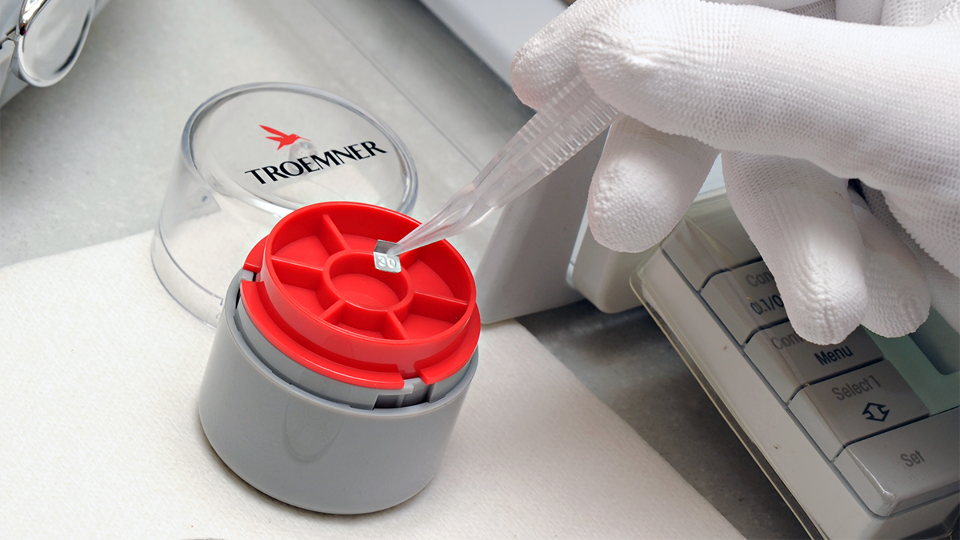 The weight case in use in a laboratory. A gloved hand is seen interacting with the precision weights and a scale.