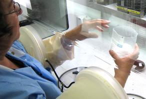 A clinician is seen with her arms through the circular arm ports of the Anaerobic Workstation while she is manipulating a glass beaker inside the device.