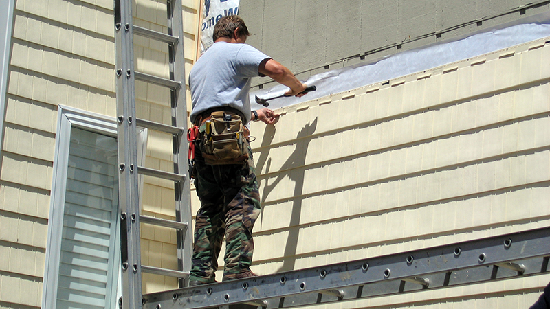 A construction worked is shown attaching cream-colored cedar shake siding to a house.