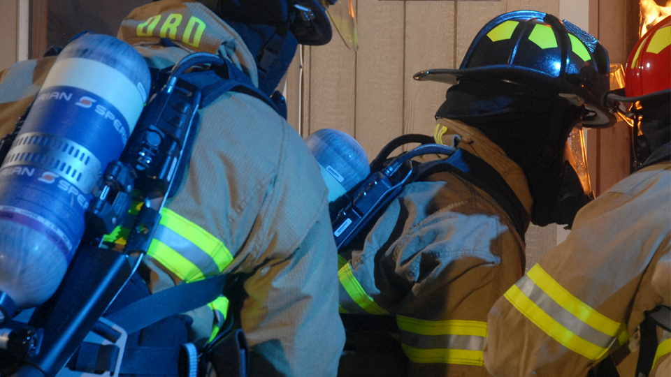 Firefighters wearing SCBA masks, moving together in a group into a burning structure.