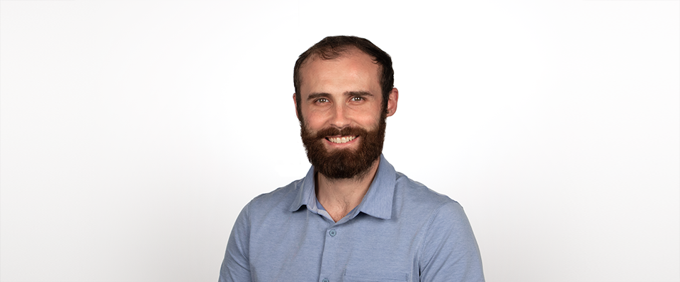 A headshot of Chris is shown. Chris is standing in front of a white background and is wearing a blue button-down shirt.