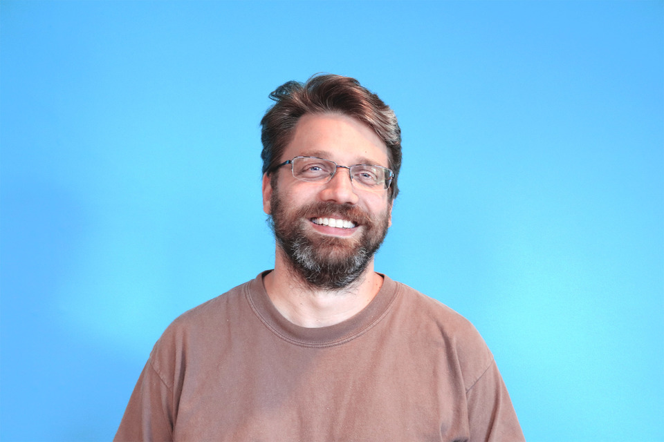 Headshot of Gregory in front of a blue wall, wearing a brown t-shirt.