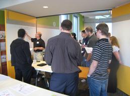 Several people are gathered around a table sharing ideas while another person records them on a whiteboard.