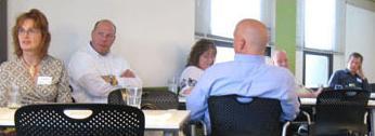 Several people sit around a table listening to a presenter give a presentation.