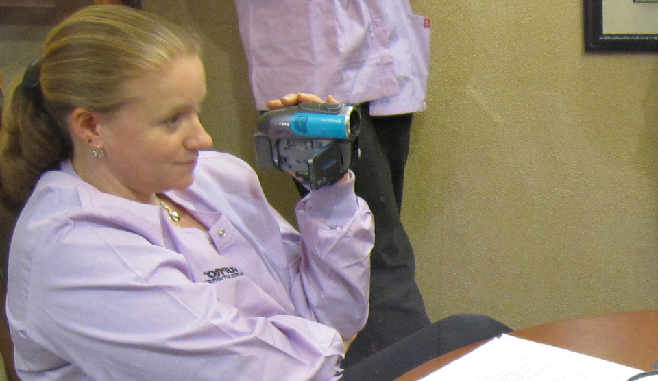 Carolynn, wearing purple scrubs, sits at a table and holds a camcorder, recording interview participants who are off-screen.
