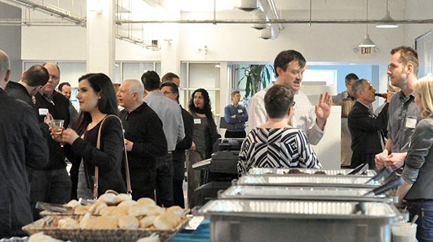 A buffet table is in the foreground, while a crowd socializes at the Daedalus office in the background.