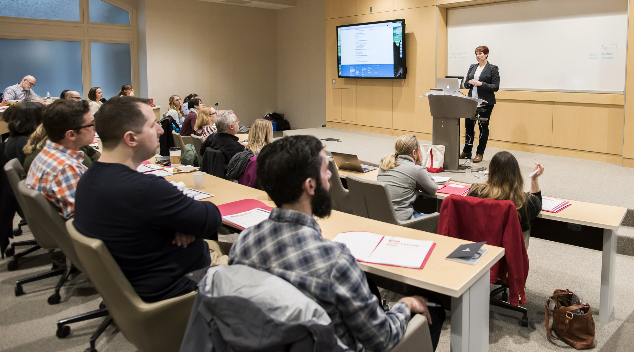 Several people are sitting in a tiered lecture hall while a woman in a black suit gives a presentation at the front of the hall.