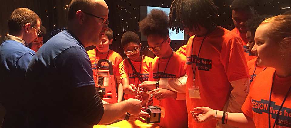 Two Daedalus employees are standing at a table with 7-8 kids attending Design Live Here. The Daedalus employees are demonstrating products developed by Daedalus, including an area gas monitor and a personal gas pump typically worn by field workers.