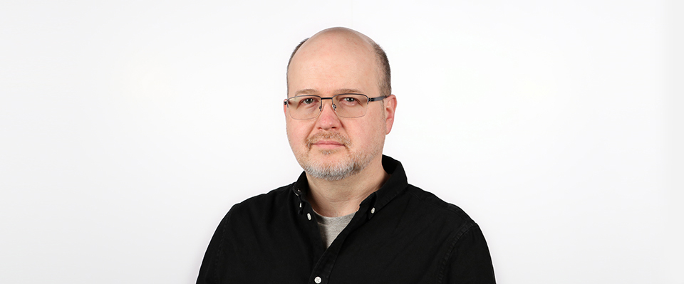 A headshot of Chris is shown on a white background. Chris is wearing glasses, a black button-down shirt, and a grey undershirt.