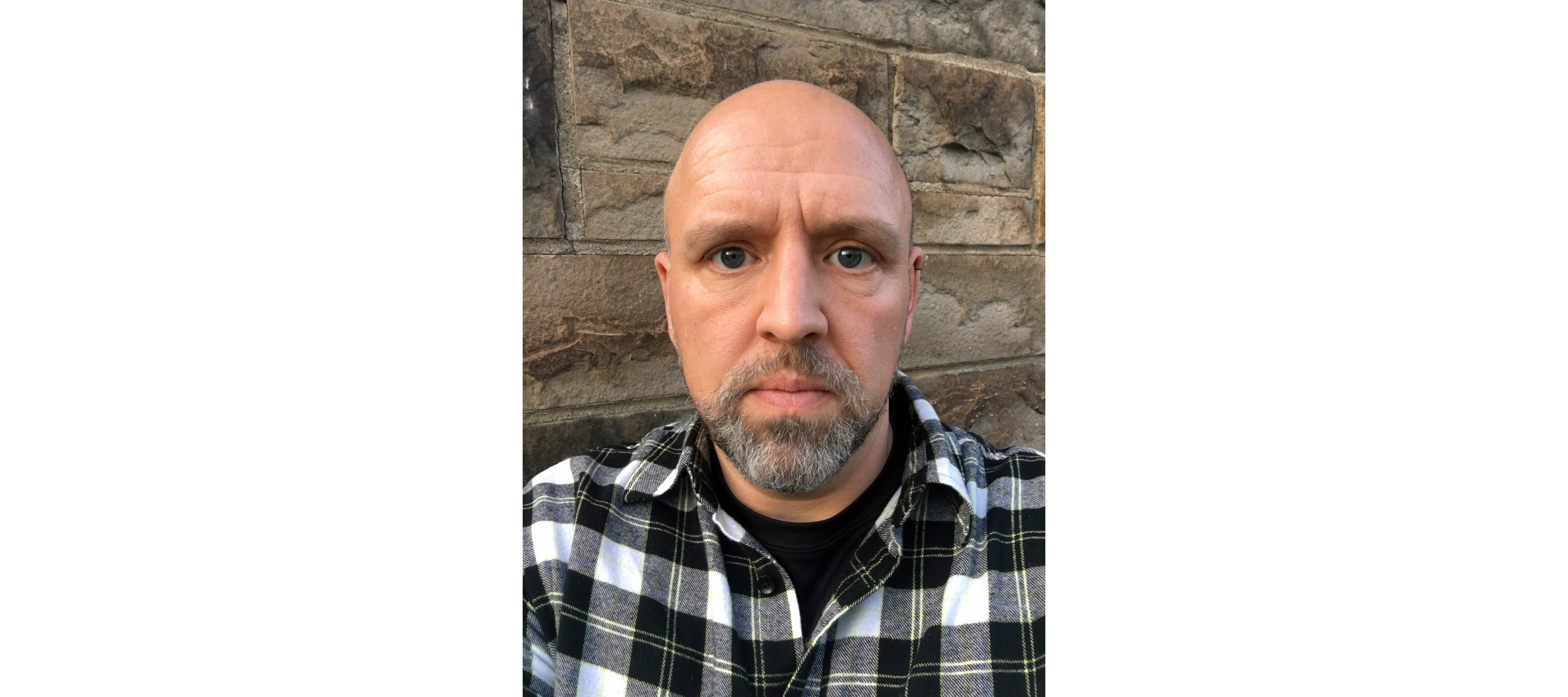 A headshot of Keith is shown. Keith is in front of a beige brick background and is wearing a black and white button-down shirt with a black t-shirt.