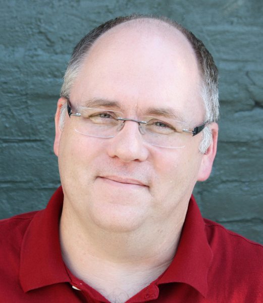 A headshot of Matt on a background of green bricks. Matt is wearing a dark red shirt and glasses.