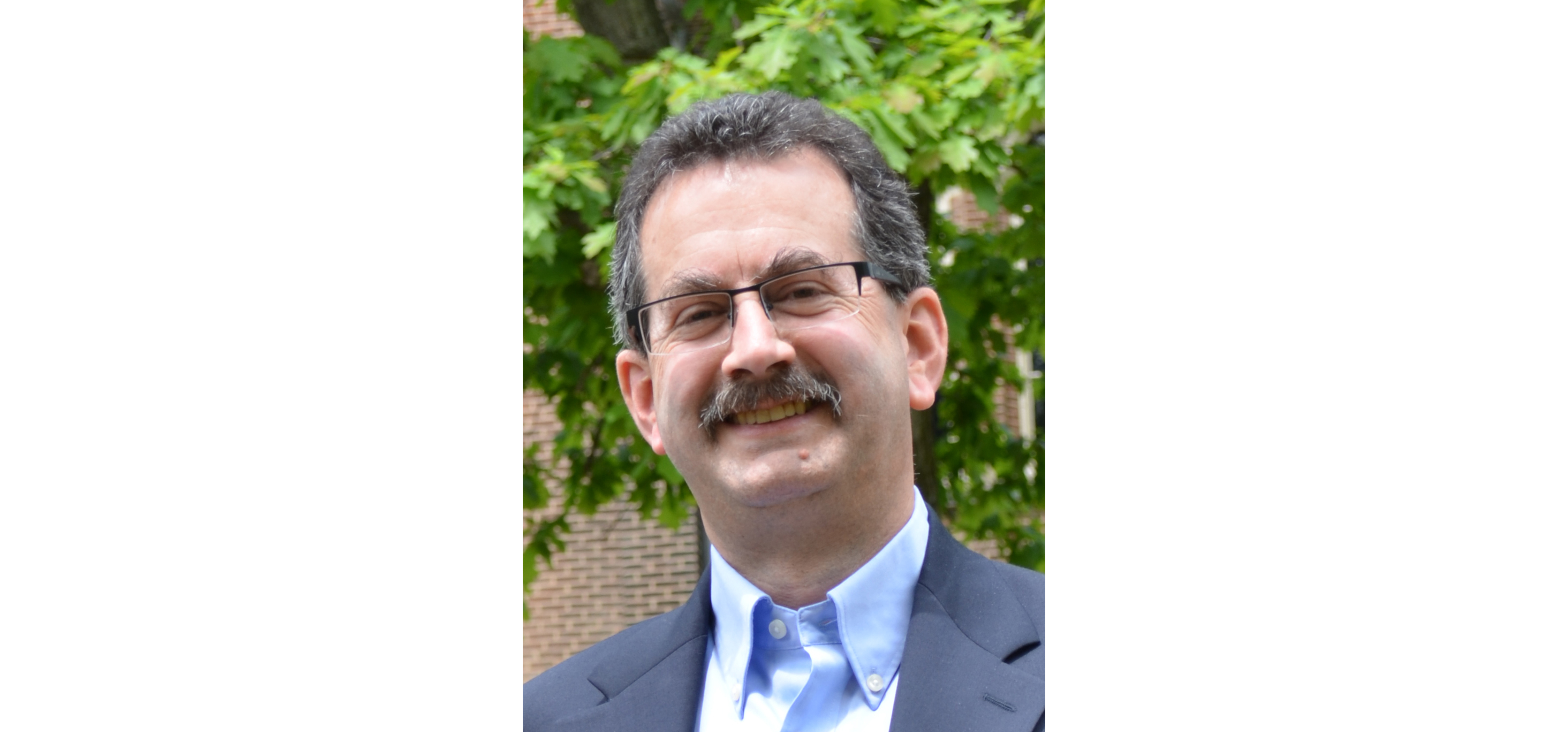 A headshot of Craig wearing a suit and tie with a tree and brick wall in the background.