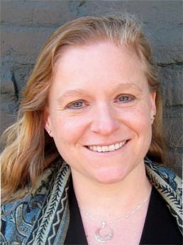 Headshot of Carolynn in front of a green brick wall, wearing a black shirt and a patterned shawl.